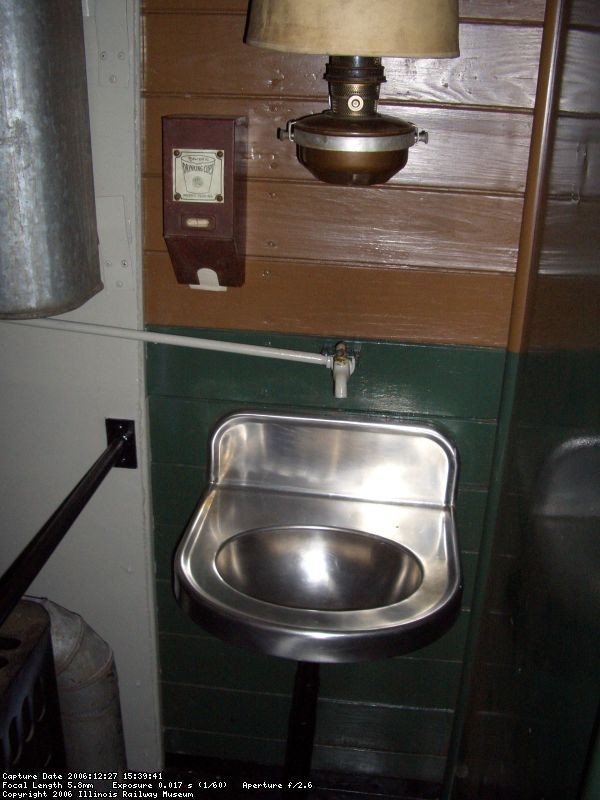 12.27.06 - THE SINK WHICH IS GRAVITY SUPPLIED BY WATER FROM THE TANK ON THE LEFT.  THE WATER TANK IS LOCATED BEHIND THE STOVE AND THE WATER WOULD BE HEATED BY THE STOVE WHEN IT WAS IN USE. THE WATER TANK IS SIGNED BY A FORMER CNW EMPLOYEE WHEN THE CAR WAS IN CHADRON, NEBRASKA. LIGHT IN THE CAR WAS FROM KEROSENE LAMPS.  PAPER CUPS ARE CONTAINED IN THE HOLDER TO THE LEFT OF THE KEROSENE LAMP AND THIS STYLE OF CUP WAS STORED IN A FLAT FORMAT.  WATER FROM THE SINK DRAINED DIRECTLY ONTO THE GROUND FROM THE DRAINPIPE.